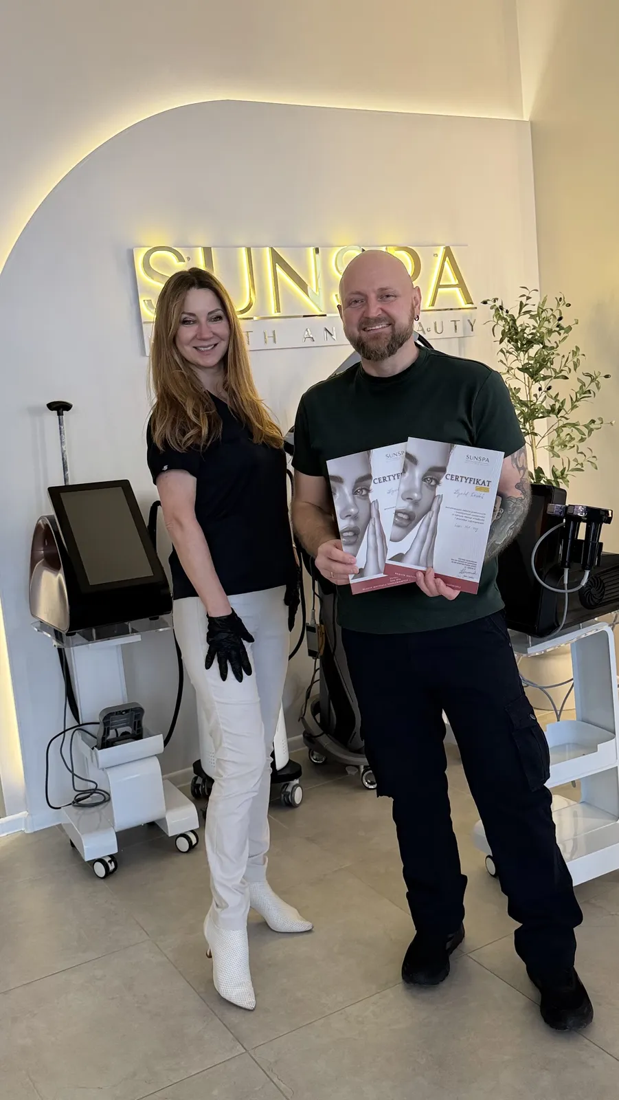 Showroom SUNSPA z podświetlanym logo na ścianie — mężczyzna z certyfikatami szkolenia obok kobiety, urządzenia kosmetyczne na wózkach